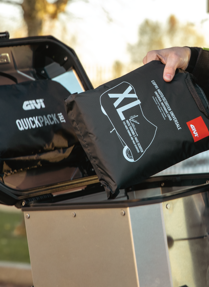 Close-up of a hand placing a rainsuit into an open GIVI top case mounted on a motorcycle.